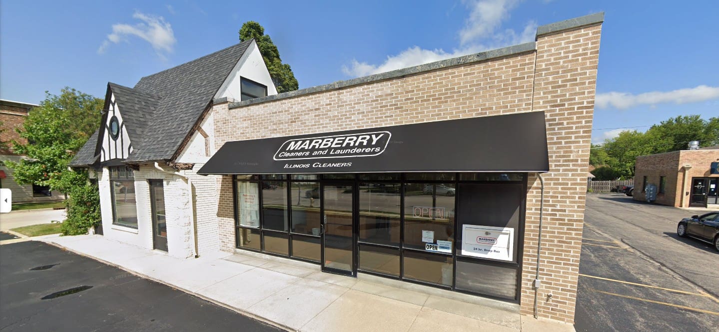 A one-story brick building with a black awning labeled "Marberry Cleaners and Launderers" and windows facing a sidewalk and parking lot under a partly cloudy sky.