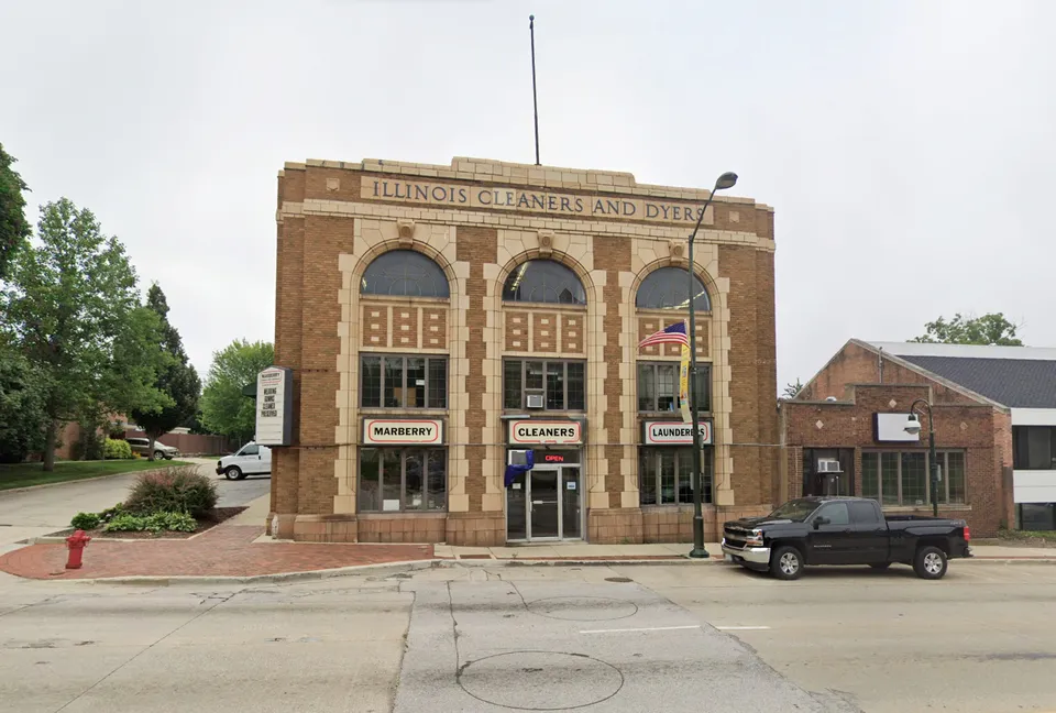A two-story brick building with arched windows labeled "Illinois Cleaners and Dyers," housing a cleaners and laundromat, with a black truck parked in front.
