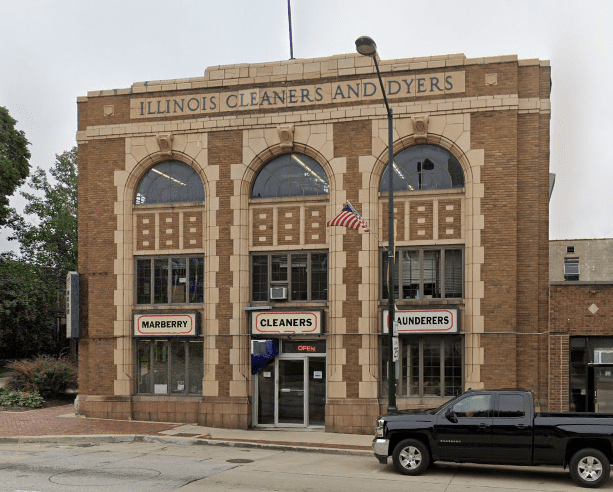 A two-story brick building labeled "Illinois Cleaners and Dyers" with three arched windows, an American flag, and a black pickup truck parked in front.