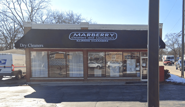 A brick storefront with a black awning reading "Marberry Cleaners and Launderers Illinois Cleaners." Large windows display various notices. A van is parked to the left.