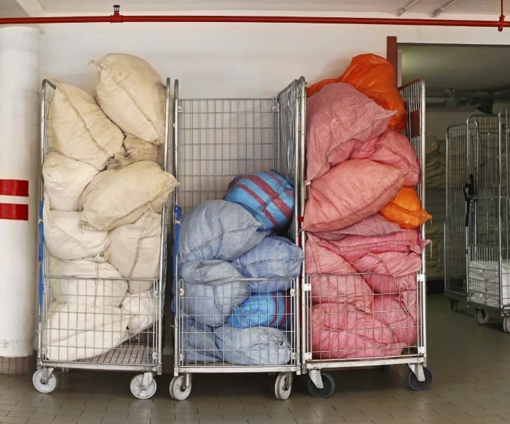 Three metal carts filled with large bags of laundry in various colors, positioned against a wall in a tiled indoor area.