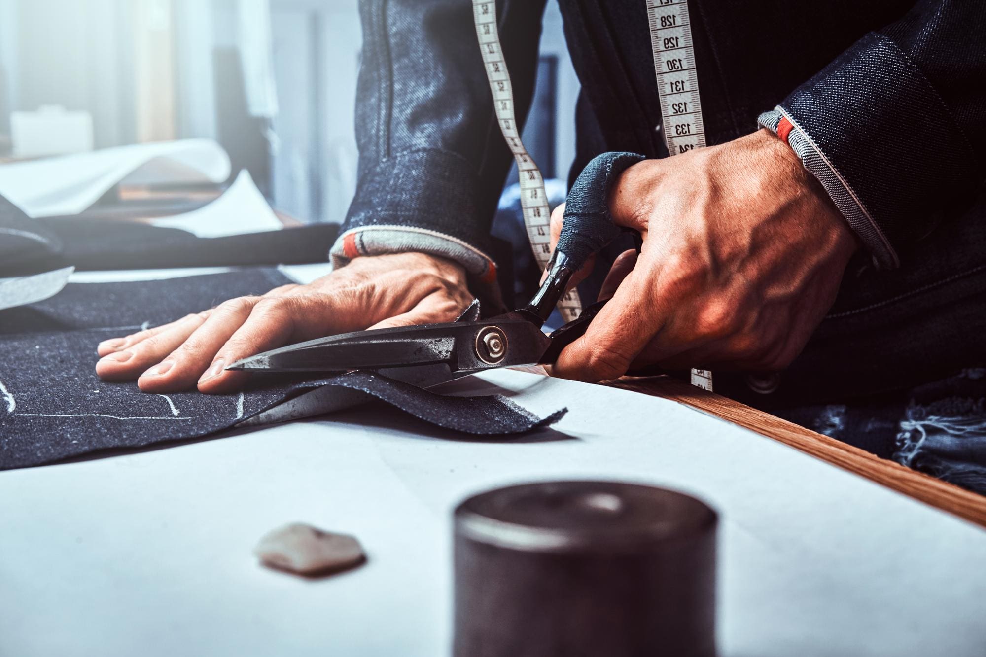 A person uses large scissors to cut fabric on a worktable, with a measuring tape draped around their neck and sewing tools nearby.