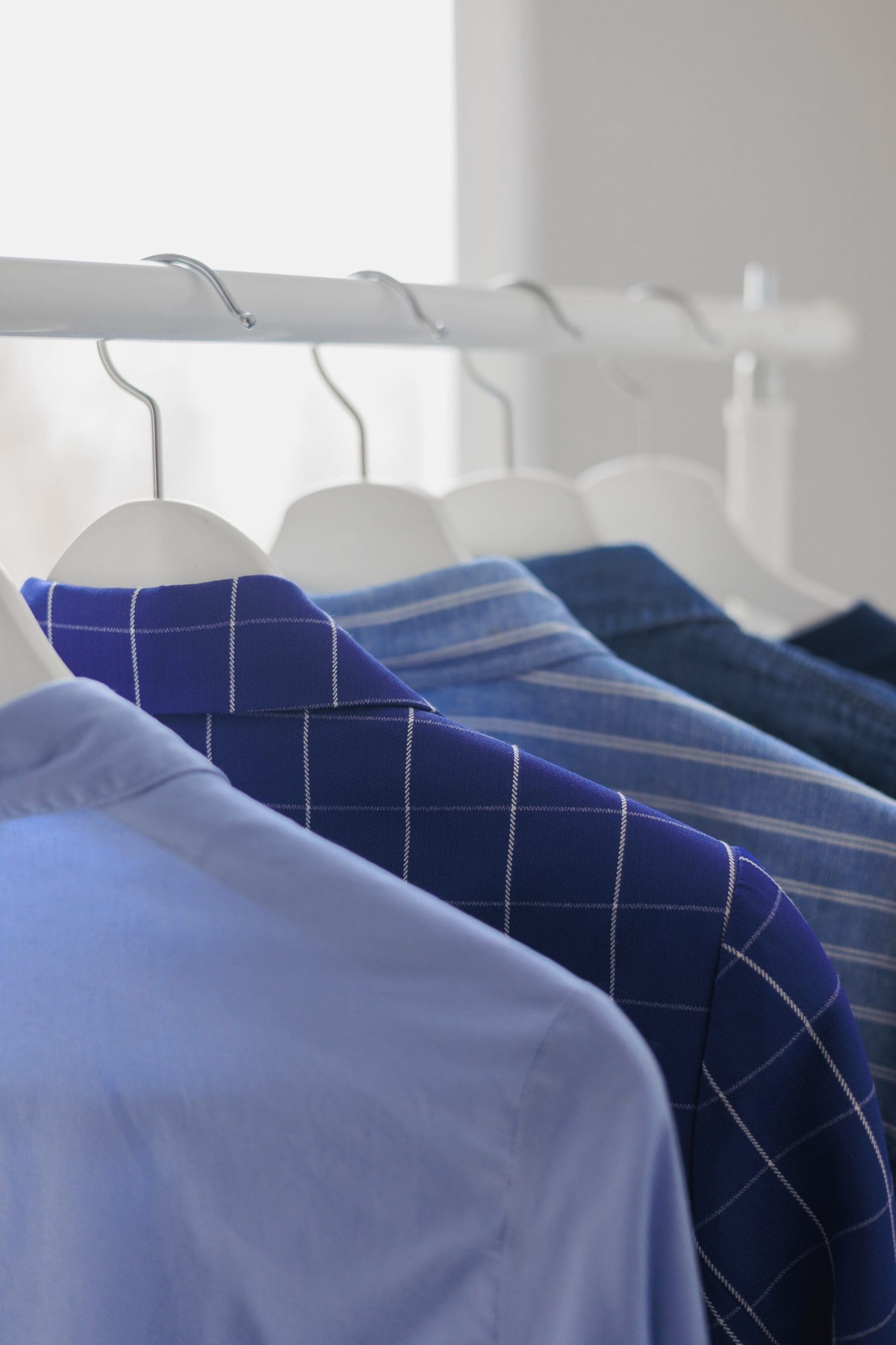 Four blue shirts with different patterns and shades are hanging on white hangers on a clothing rack, with a blurred window in the background.