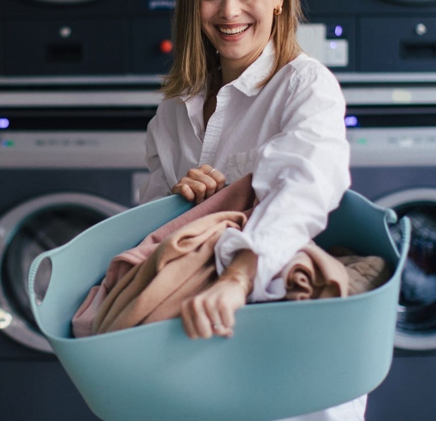 A person wearing a white shirt holds a large blue laundry basket filled with clothes in front of washing machines at a laundromat.