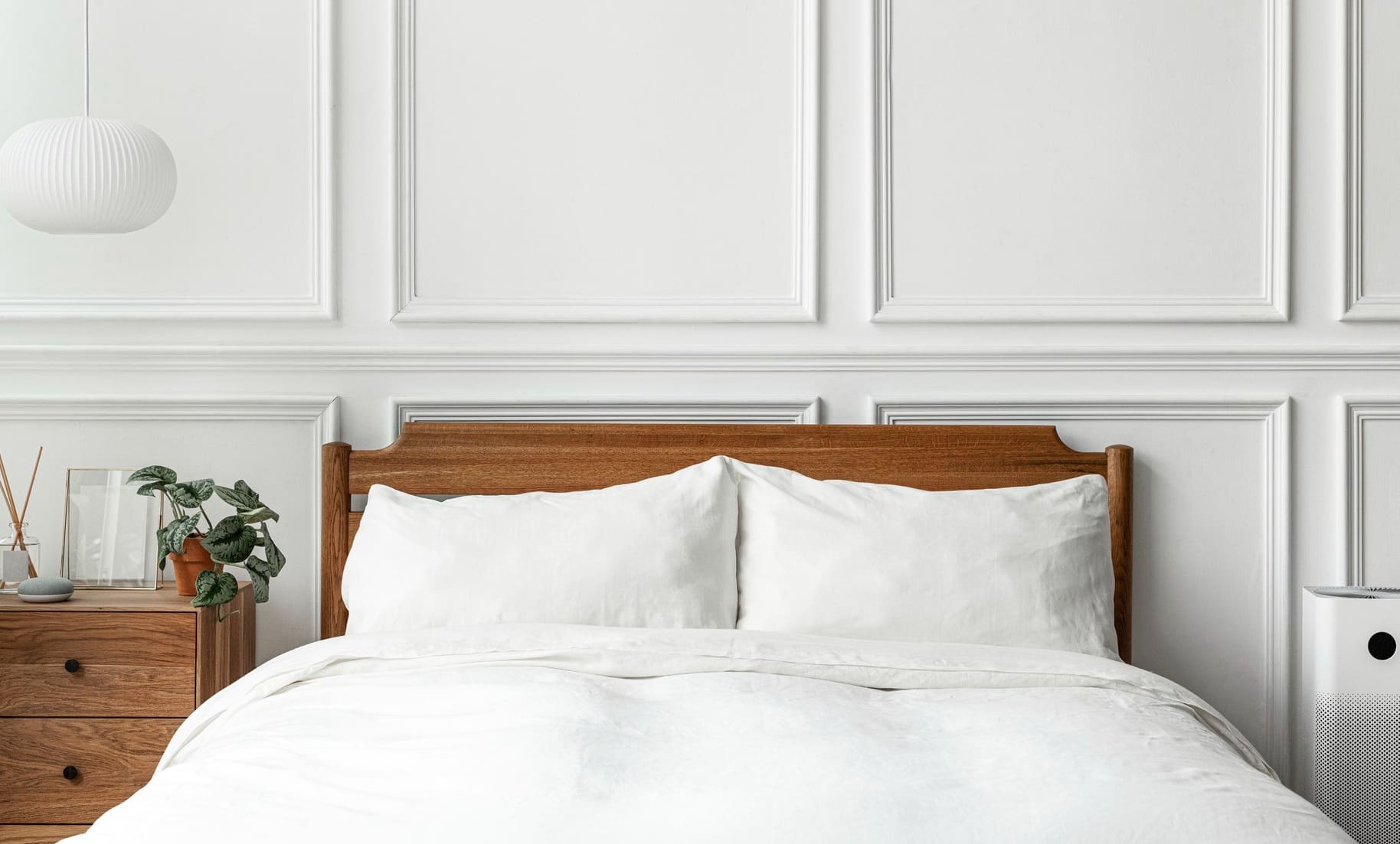A neatly made bed with white bedding and a wooden headboard is centered against a white paneled wall, with a nightstand and a plant on the left.