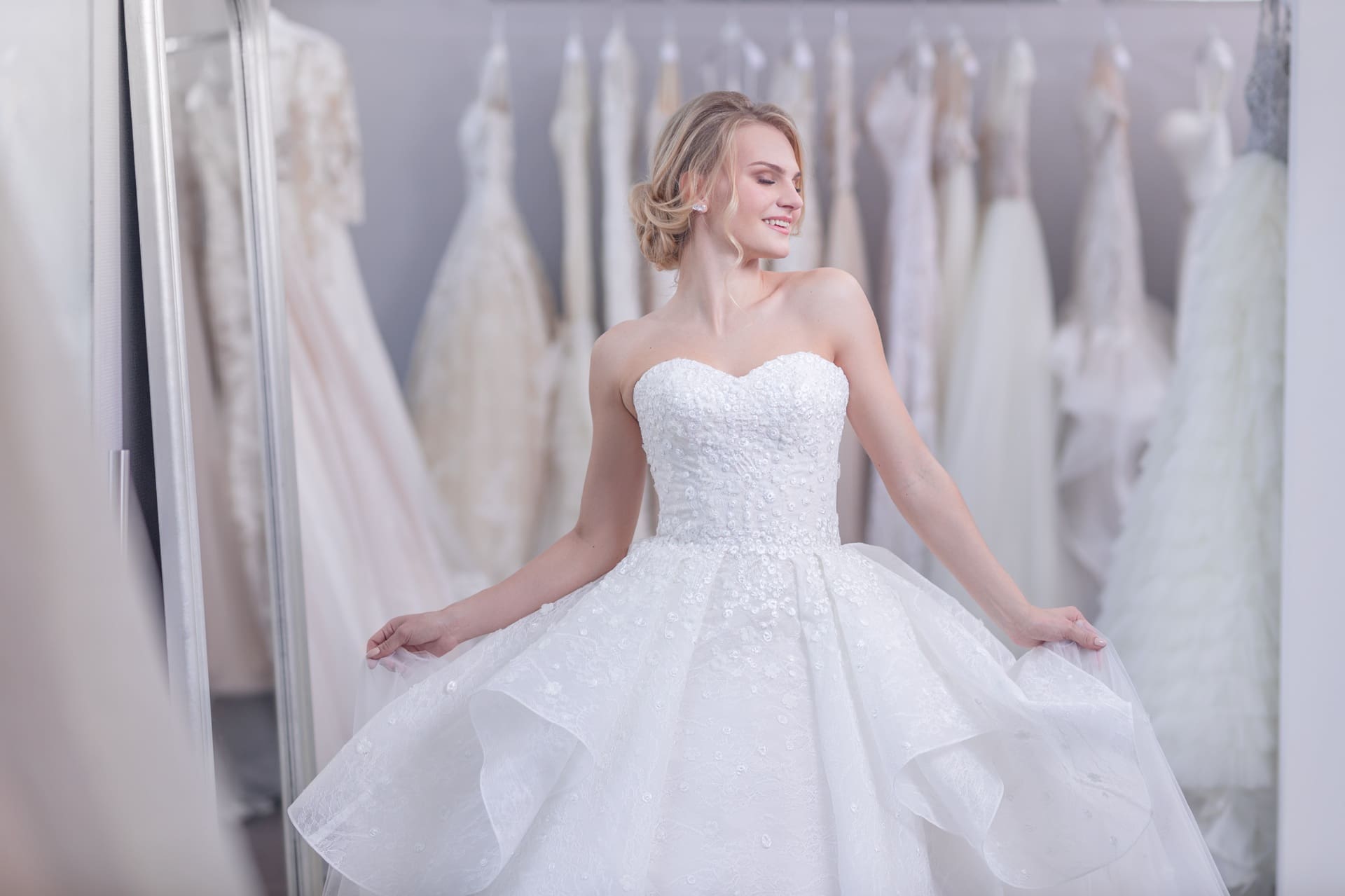 Woman wearing a strapless white wedding dress stands in front of a mirror in a bridal shop, surrounded by other wedding gowns on hangers.