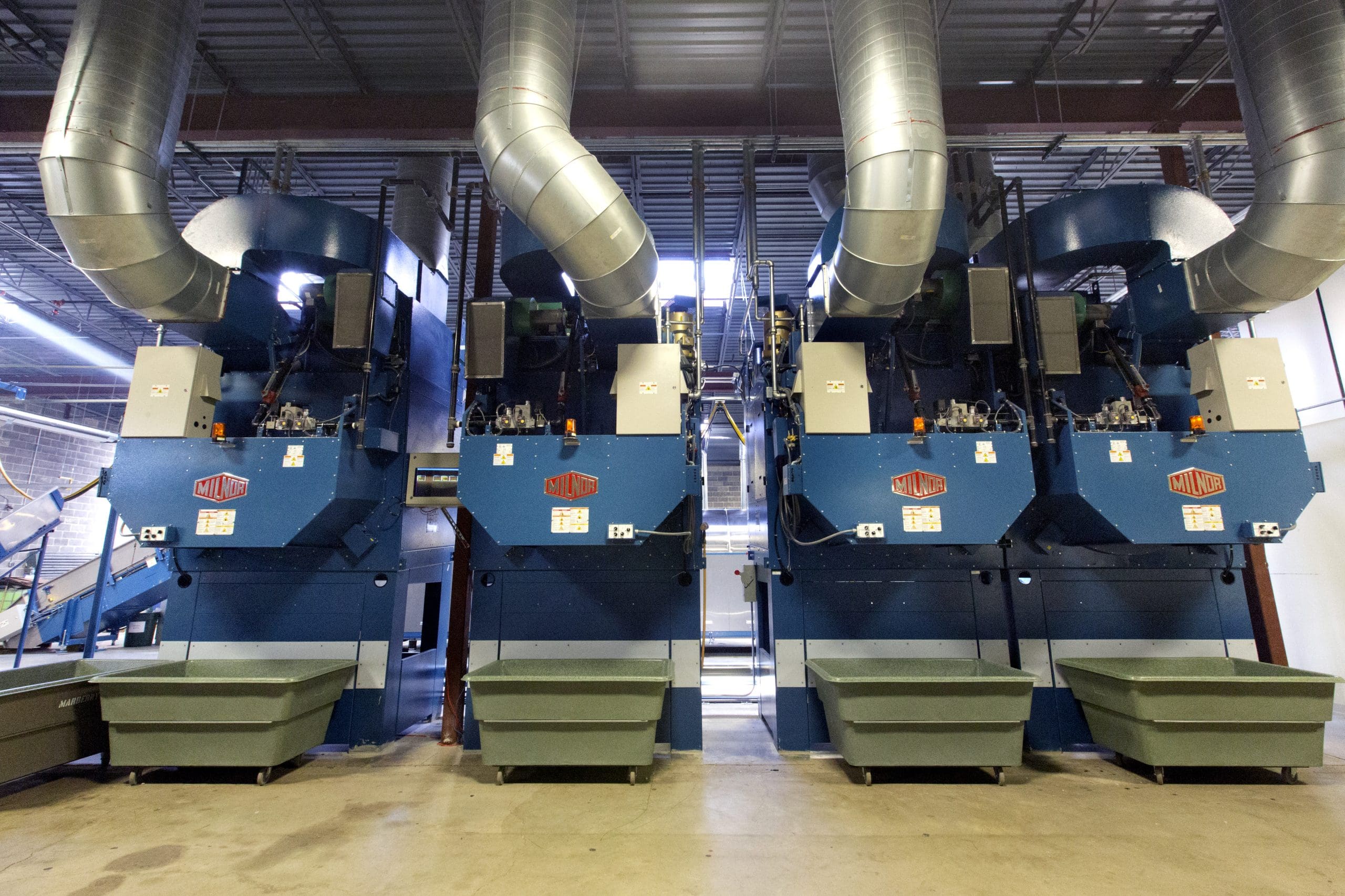Three large industrial machines with ducts overhead are lined up side by side in a spacious factory setting.