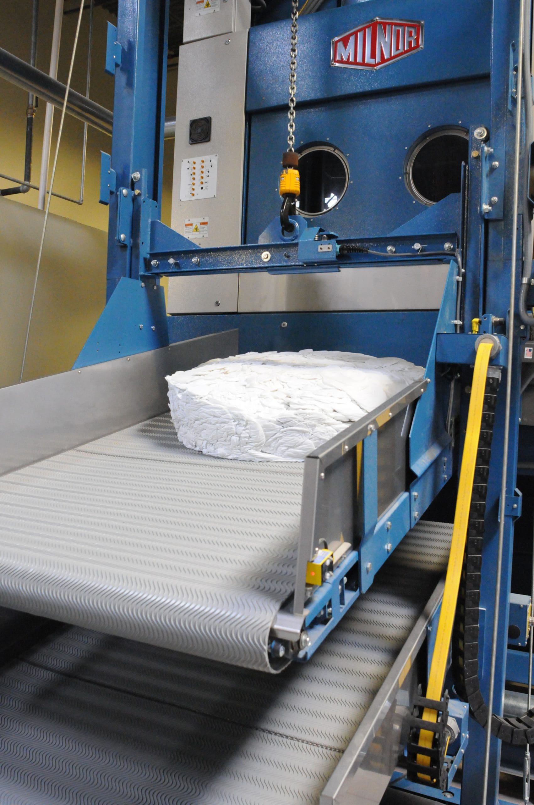 A large industrial washing machine unloads a stack of white linens onto a conveyor belt in a commercial laundry facility.