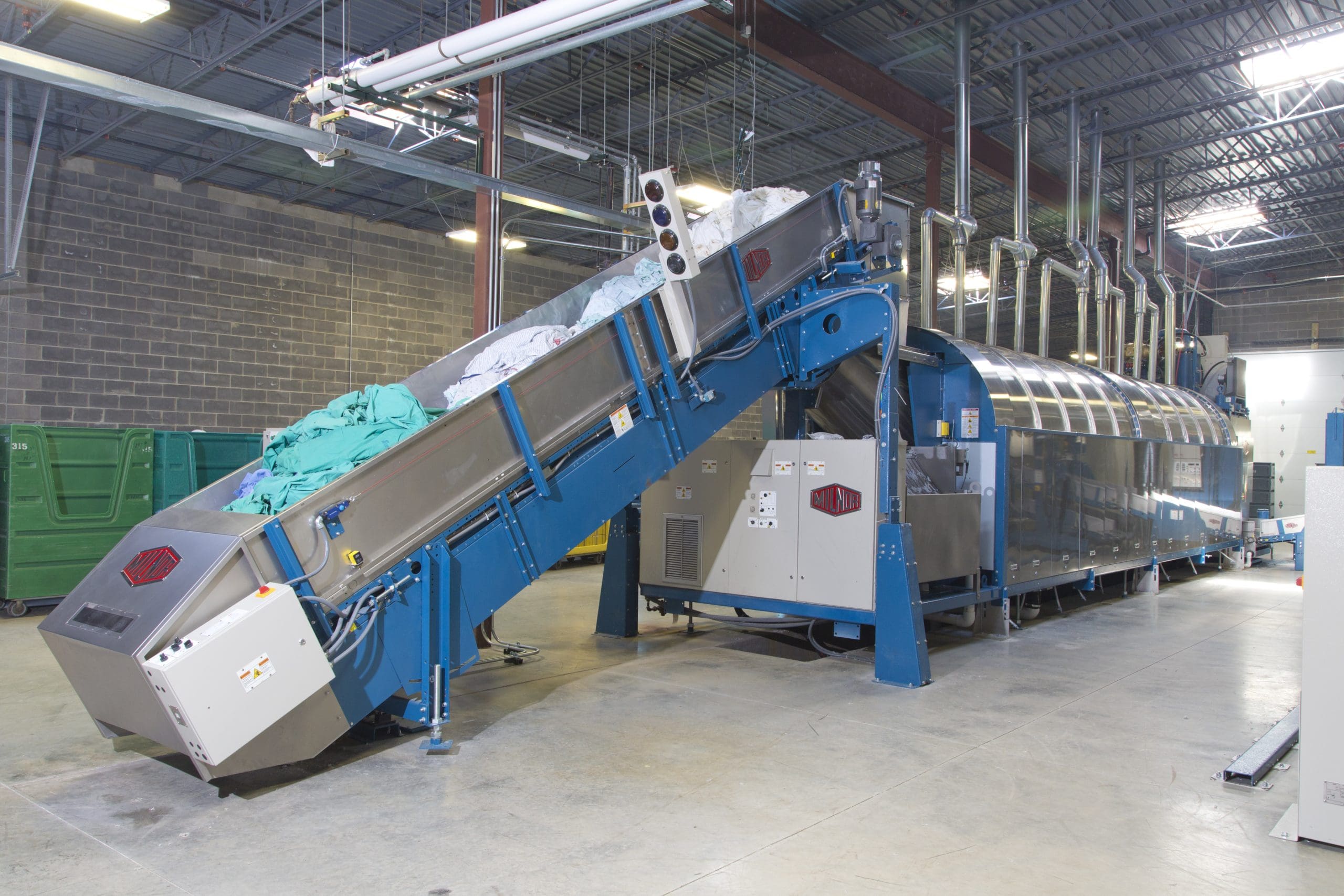 Large industrial washing machine and conveyor system processing laundry in a spacious facility with concrete floors and exposed ceilings.