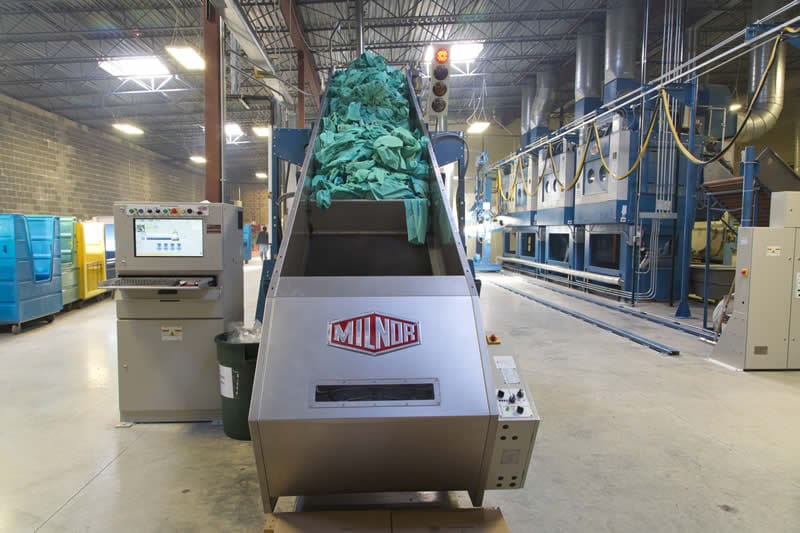 A commercial laundry machine loaded with green linens is situated in an industrial facility, with control panels and industrial equipment visible in the background.
