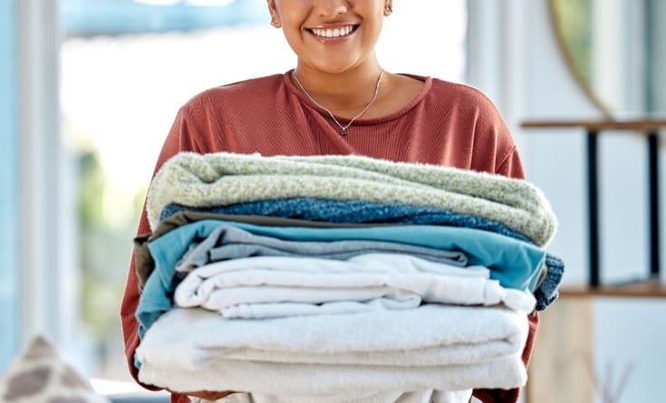 A person smiles while holding a neatly folded stack of towels and clothes indoors.