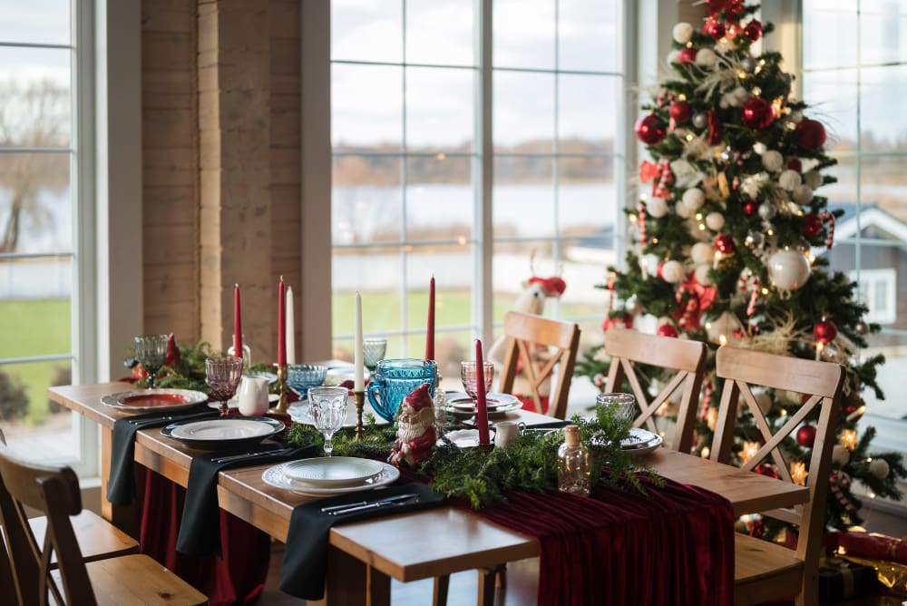 A decorated dining table set for a meal with red and green accents, candles, and festive greenery, next to a Christmas tree by large windows.