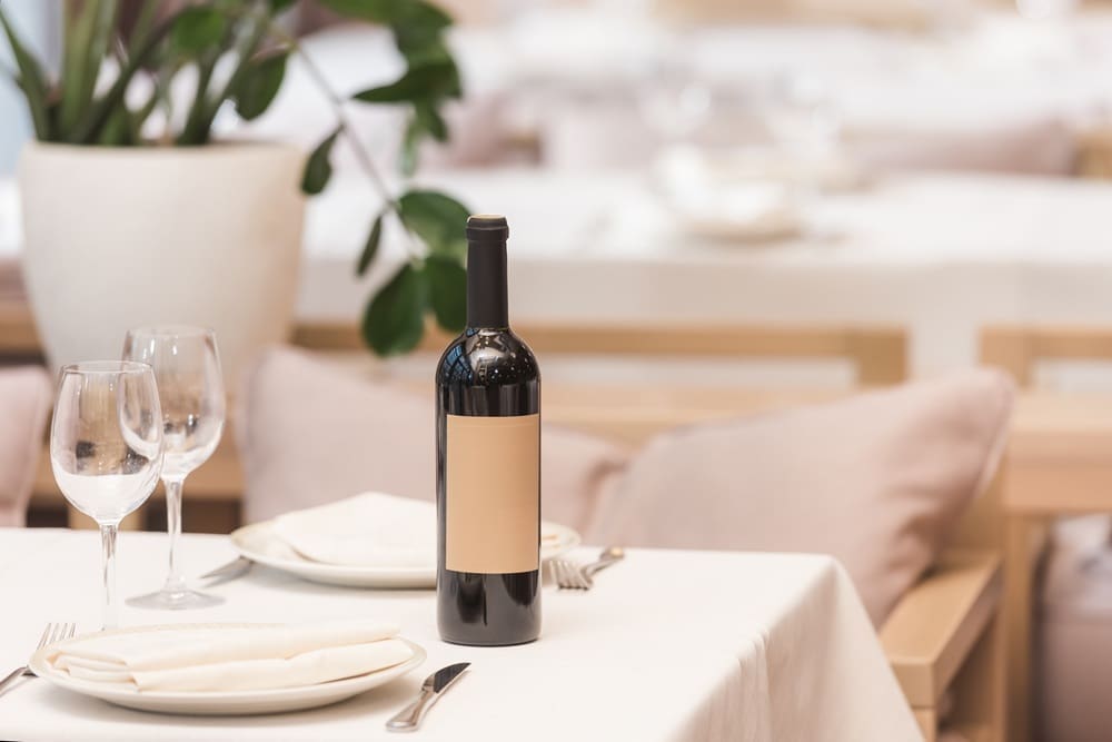 A bottle of wine with a blank label sits on a set dining table with two wine glasses, plates, cutlery, and a potted plant in the background.