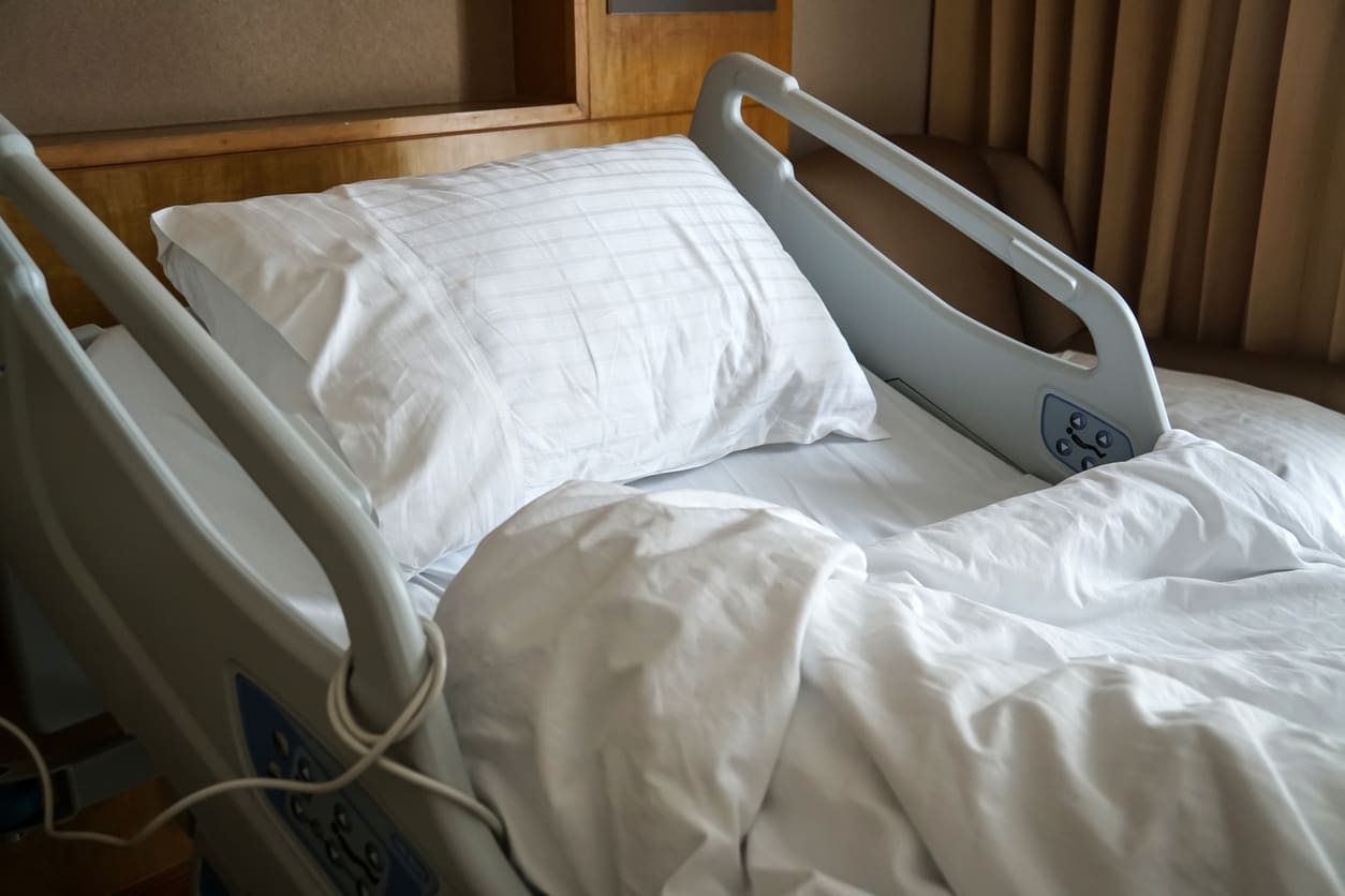 A neatly made hospital bed with white sheets and pillow in a patient room, featuring adjustable side rails and a wooden headboard.