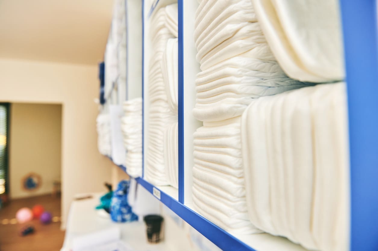 Stacks of folded white diapers are neatly arranged in blue cubby shelves, with a room and scattered toys visible in the background.