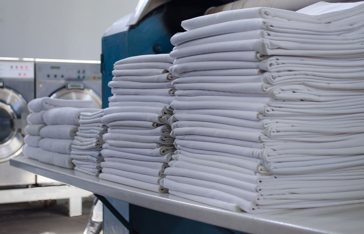 Stacks of neatly folded white towels and linens on a table in a laundromat, with industrial washing machines in the background.