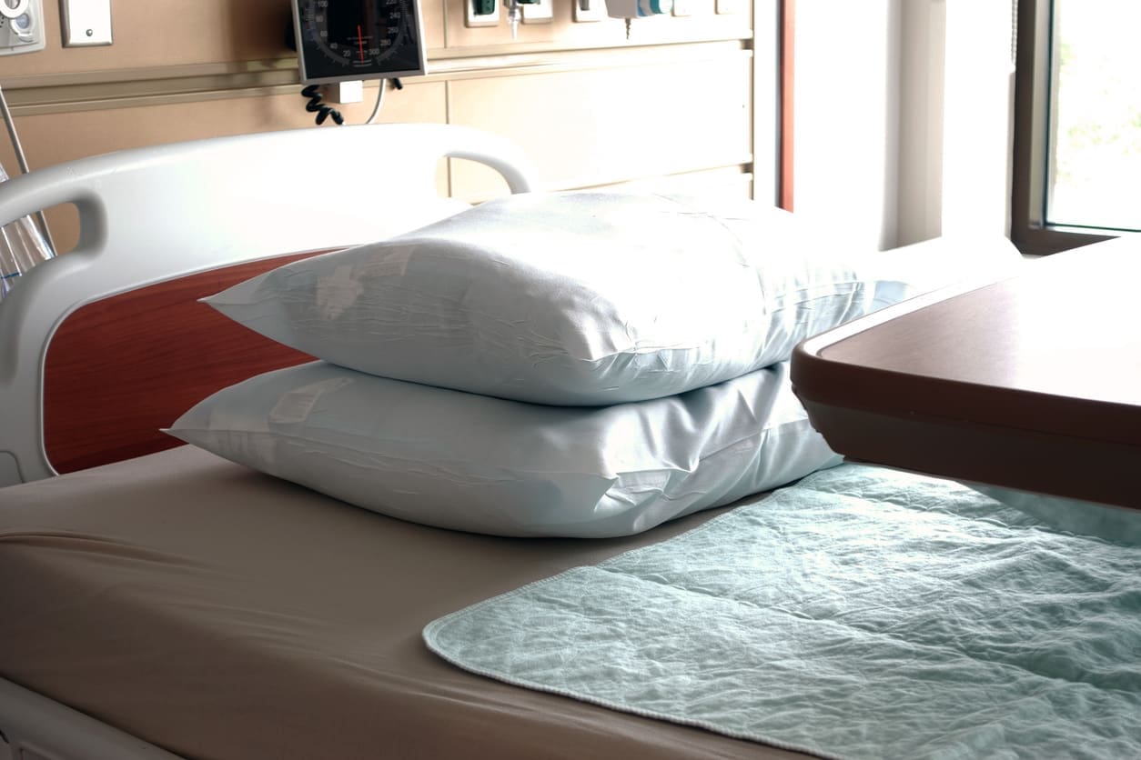 Hospital bed with two stacked pillows, a bedside tray, and medical equipment in the background. Natural light comes through a nearby window.
