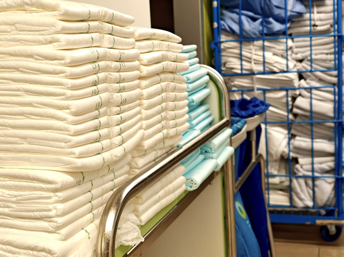 Stacks of folded white and light blue linens are arranged on a cart, with more folded linens organized in a blue wire storage rack in the background.