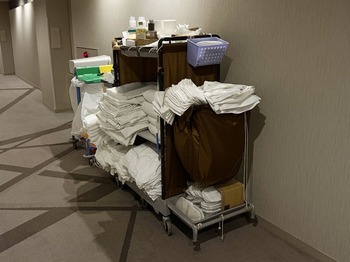 A hotel housekeeping cart in a hallway, stacked with folded white linens, cleaning supplies, waste bags, and other maintenance items.