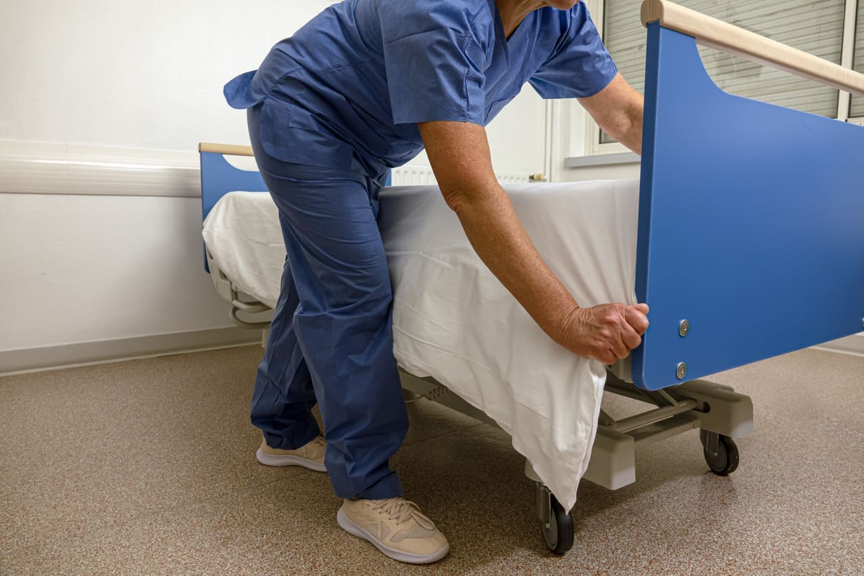Healthcare worker in blue scrubs tucks a white sheet under the mattress of a hospital bed in a clean, well-lit room.