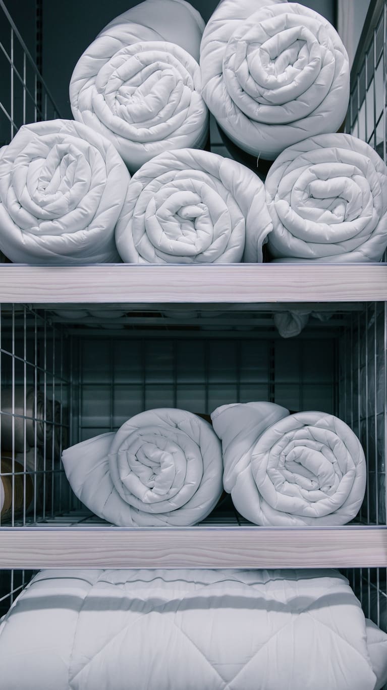 Rolled white comforters are neatly stacked on two metal wire shelves, with an additional folded comforter placed on the bottom shelf.