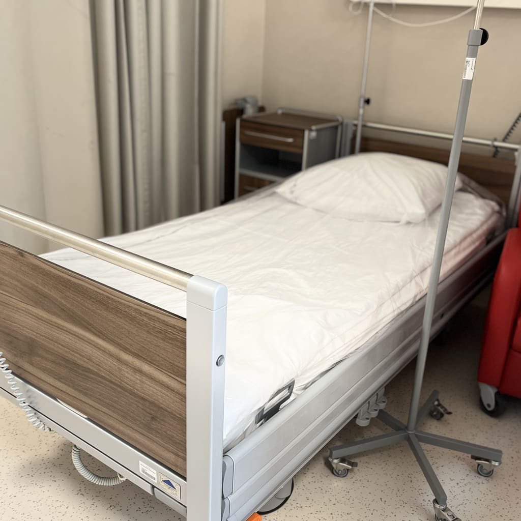 A hospital room with an empty bed, white bedding, an IV stand, bedside table, and a red chair, all set on a beige tiled floor.