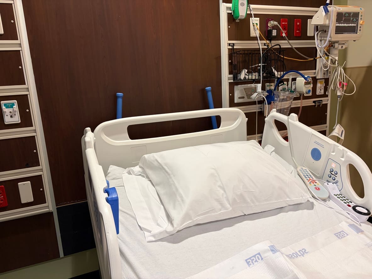 A hospital bed with white sheets and pillow, surrounded by medical equipment, monitors, a nurse call button, and wall outlets in a patient room.