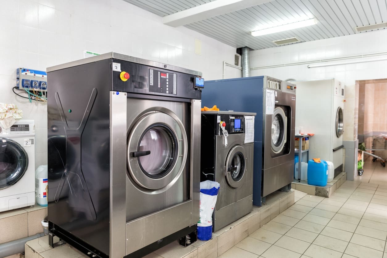 Row of large industrial washing machines in a laundry room with tiled floors and cleaning supplies visible nearby.