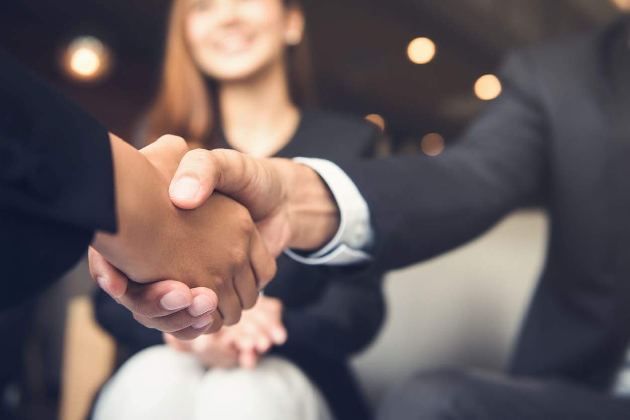 Two people in business attire shake hands in the foreground, while another person sits in the background, slightly out of focus.